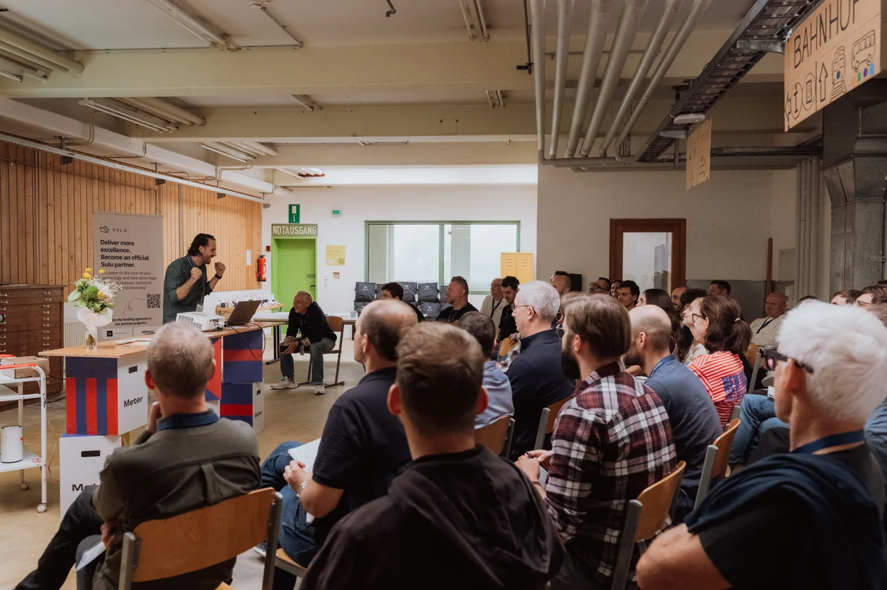 A speaker presents to an engaged audience at a Sulu community event in an industrial-style venue. The collaborative setting brings together agencies, developers, and digital teams to exchange practical insights about flexible content management and web development. Sulu community presentation